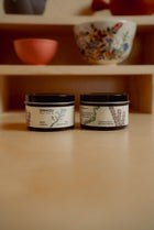 Two containers with labels on a wooden surface, with a blurred background of shelves.